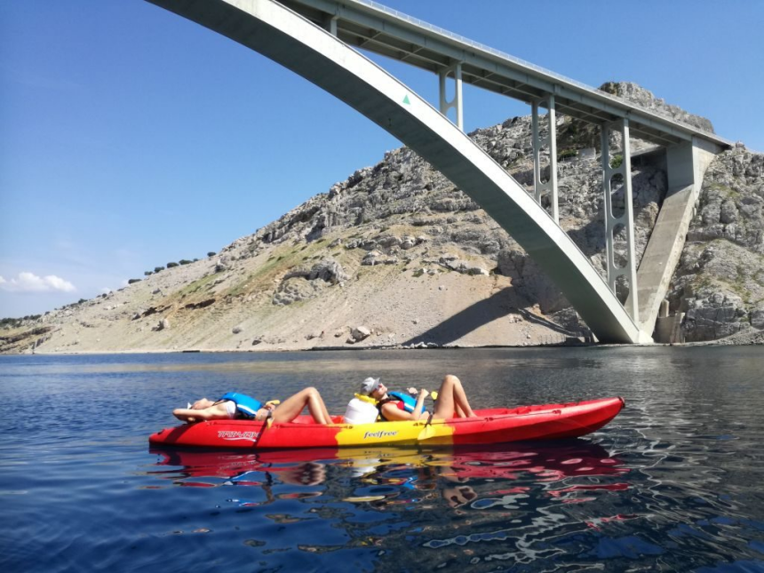 Two people relaxing on a red kayak under the Krk Bridge on a sunny day